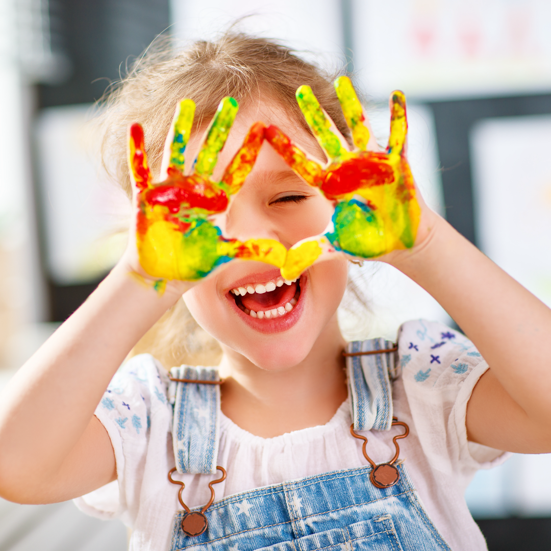 Smiling young girl with colourful painted hands held up to her face in a playful gesture, wearing denim overalls and a white shirt with blue accents, in a bright and creative indoor environment.